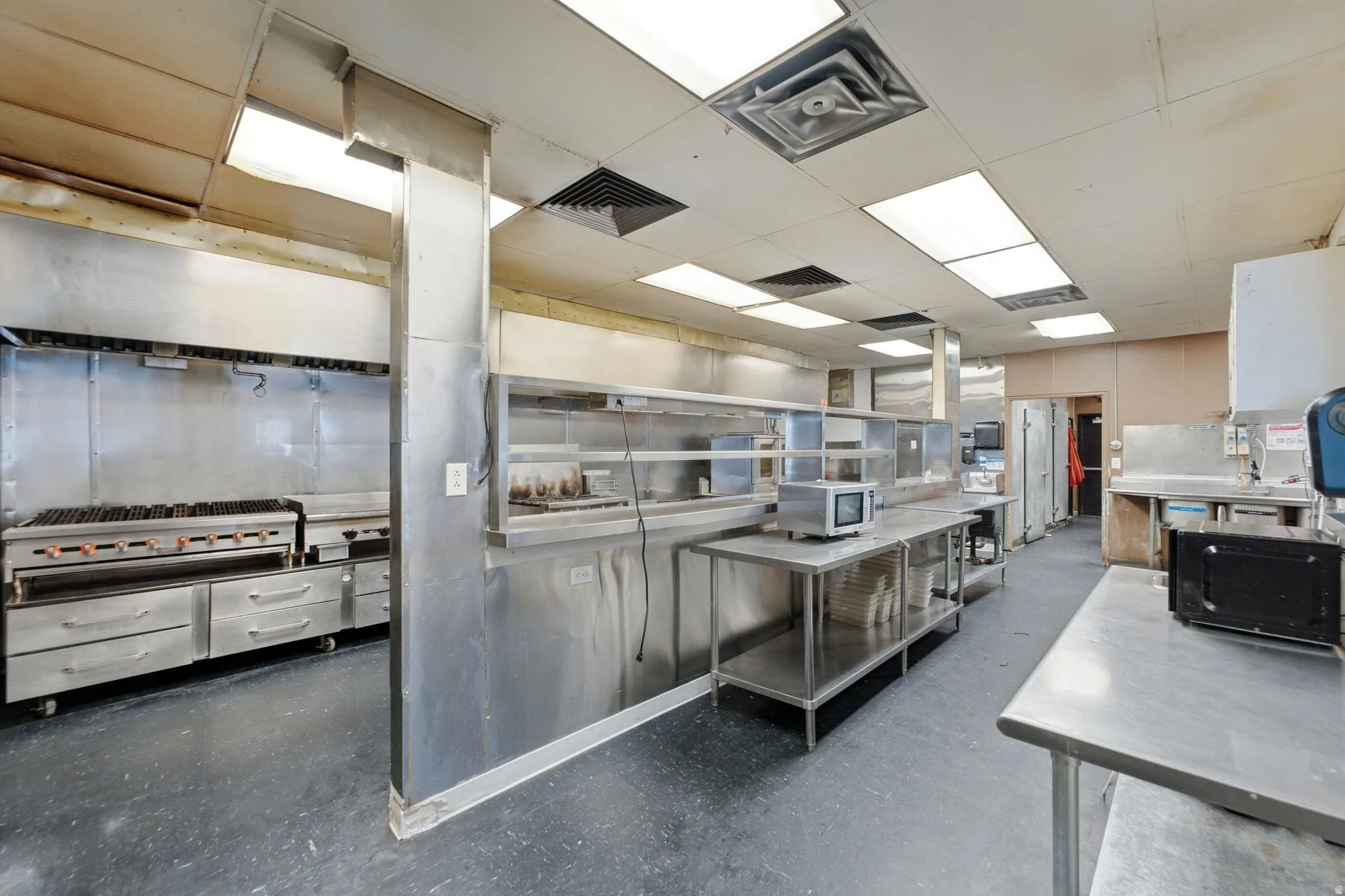 Professional-grade commercial kitchen featuring stainless steel prep and cooking stations, extensive under-counter storage, overhead ventilation systems, and durable epoxy flooring throughout