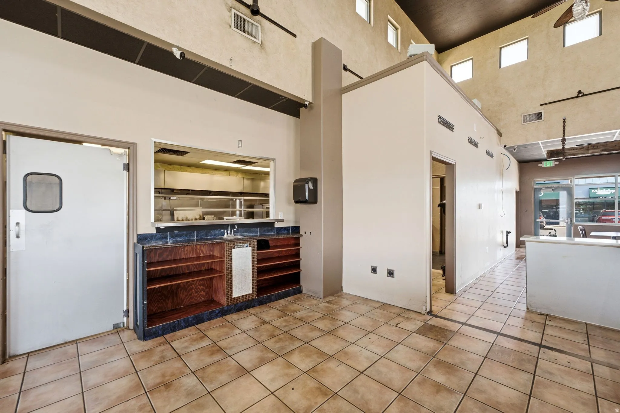 Spacious commercial kitchen featuring stainless steel pass-through window, wood-finish cabinetry with ample storage, tile flooring, high ceilings with skylights, and direct access to service entrance and retail storefront