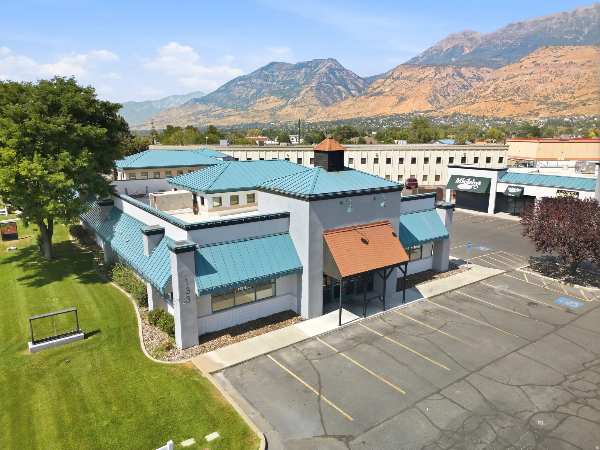 Modern commercial or mixed-use facility featuring turquoise metal roofing, clean geometric architecture with multi-level construction, expansive paved parking area, and manicured landscaping with mature trees