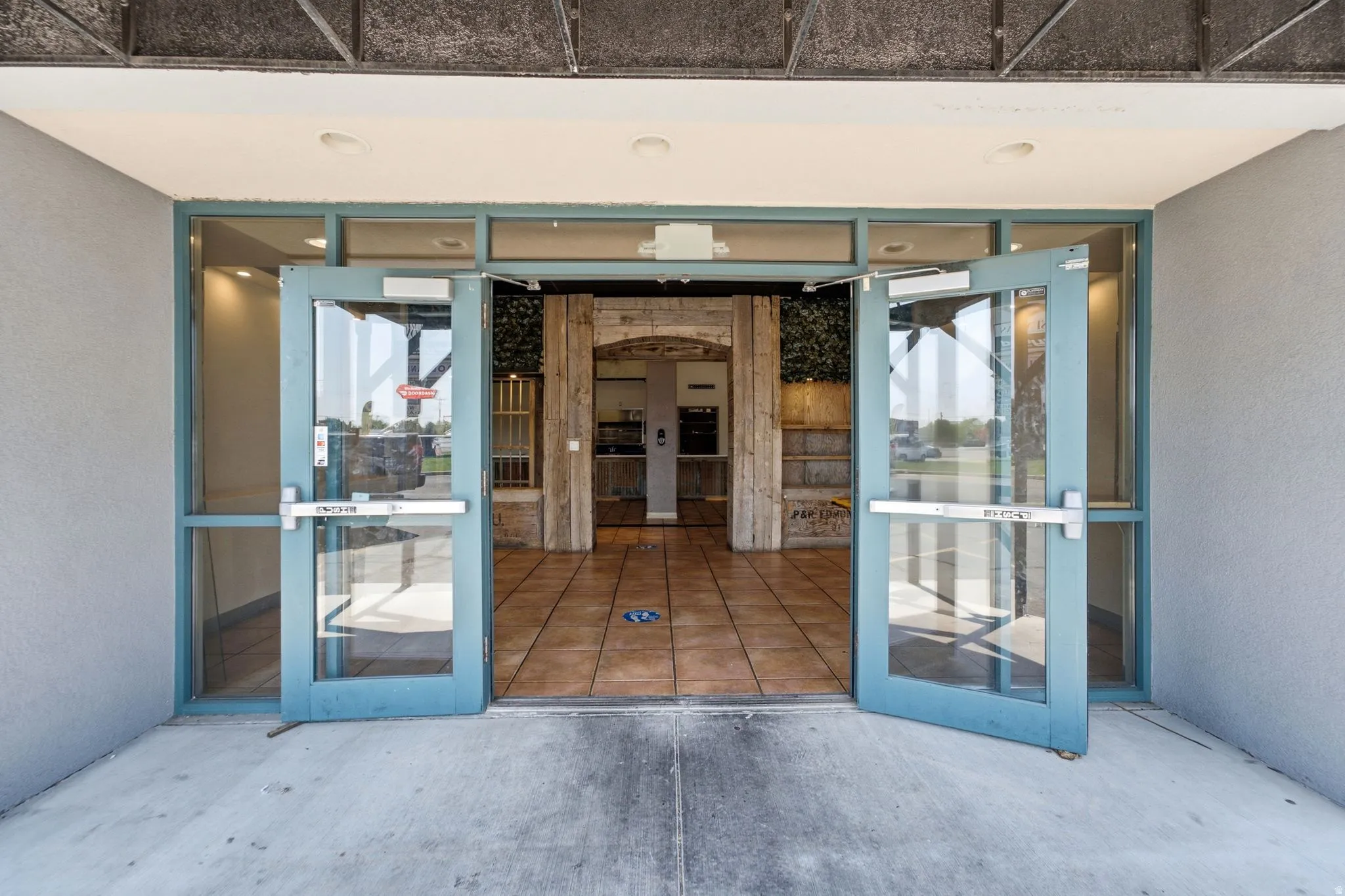 Welcoming entrance featuring expansive glass double doors with blue-framed aluminum frames, terracotta tile flooring, and sightlines through to an interior courtyard with wooden architectural detailing and natural light