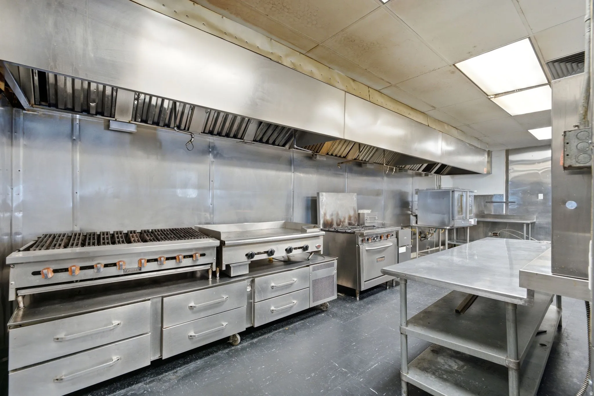 Commercial kitchen featuring stainless steel surfaces, multiple cooking stations, built-in ventilation hood, and fluorescent overhead lighting