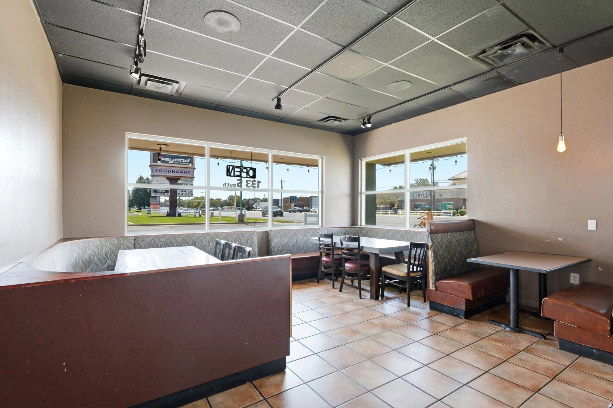 Bright, open commercial or retail space featuring expansive front-facing windows with natural light, ceramic tile flooring, drop ceiling with recessed lighting, and multiple functional zones for display or seating