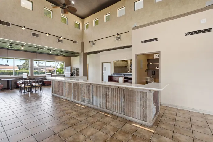 Spacious commercial or hospitality interior featuring a prominent wood-finish service counter with corrugated metal paneling, expansive tile flooring, high ceilings with clerestory windows and track lighting, and multiple service windows with wood-frame o