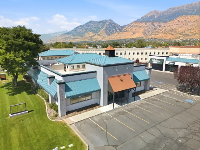 Modern commercial or mixed-use facility featuring turquoise metal roofing, clean geometric architecture with multi-level construction, expansive paved parking area, and manicured landscaping with mature trees