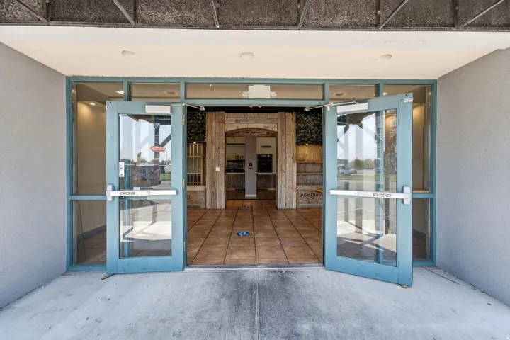 Welcoming entrance featuring expansive glass double doors with blue-framed aluminum frames, terracotta tile flooring, and sightlines through to an interior courtyard with wooden architectural detailing and natural light