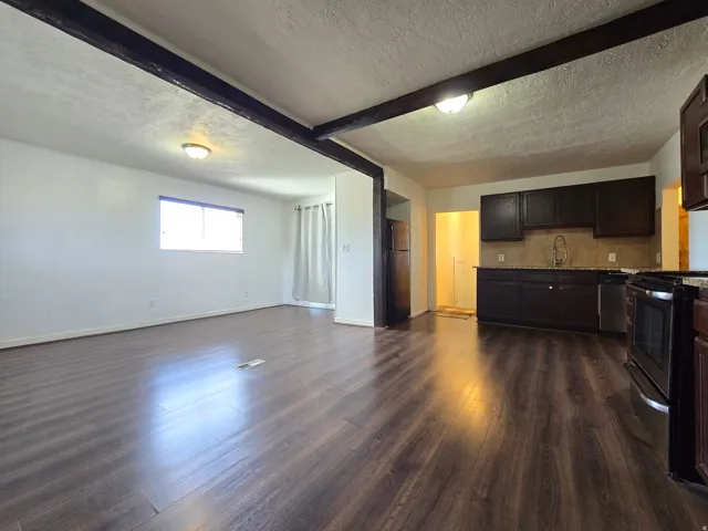 Wood-finish flooring, exposed ceiling beams, a kitchen with dark cabinetry, granite-style countertops, and a full backsplash