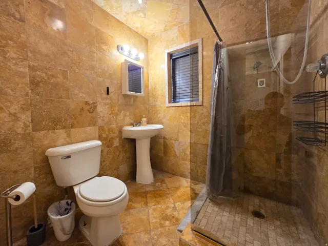 Stone-tiled walls and flooring, pedestal sink, shower enclosure with glass door, small window with blinds, and vanity light fixture