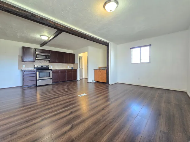 Open concept living area featuring wood-finish flooring, exposed ceiling beams, and a kitchen with stainless steel appliances, dark wood-finish cabinetry, and a neutral tile backsplash