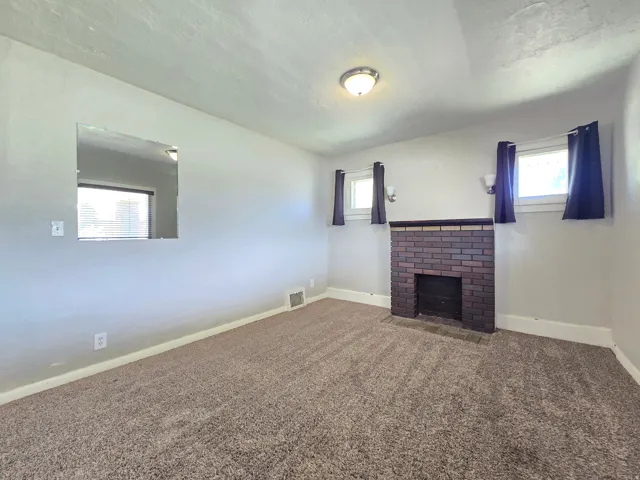 Carpeted room featuring a brick fireplace with a dark wood mantel, two windows, and a ceiling-mounted light fixture