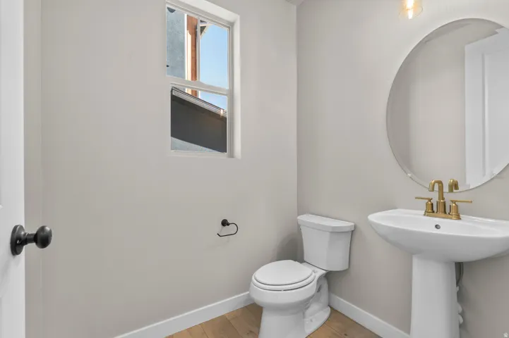 Contemporary bathroom featuring wood-finish flooring, a white pedestal sink with brass-finish fixtures, a circular mirror, and a white toilet