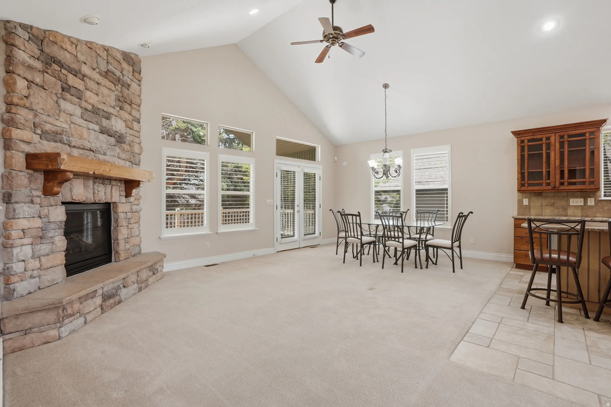 Vaulted ceiling with a ceiling fan, stone fireplace with a wood mantle, numerous windows with white blinds, and a built-in wet bar with wood cabinetry