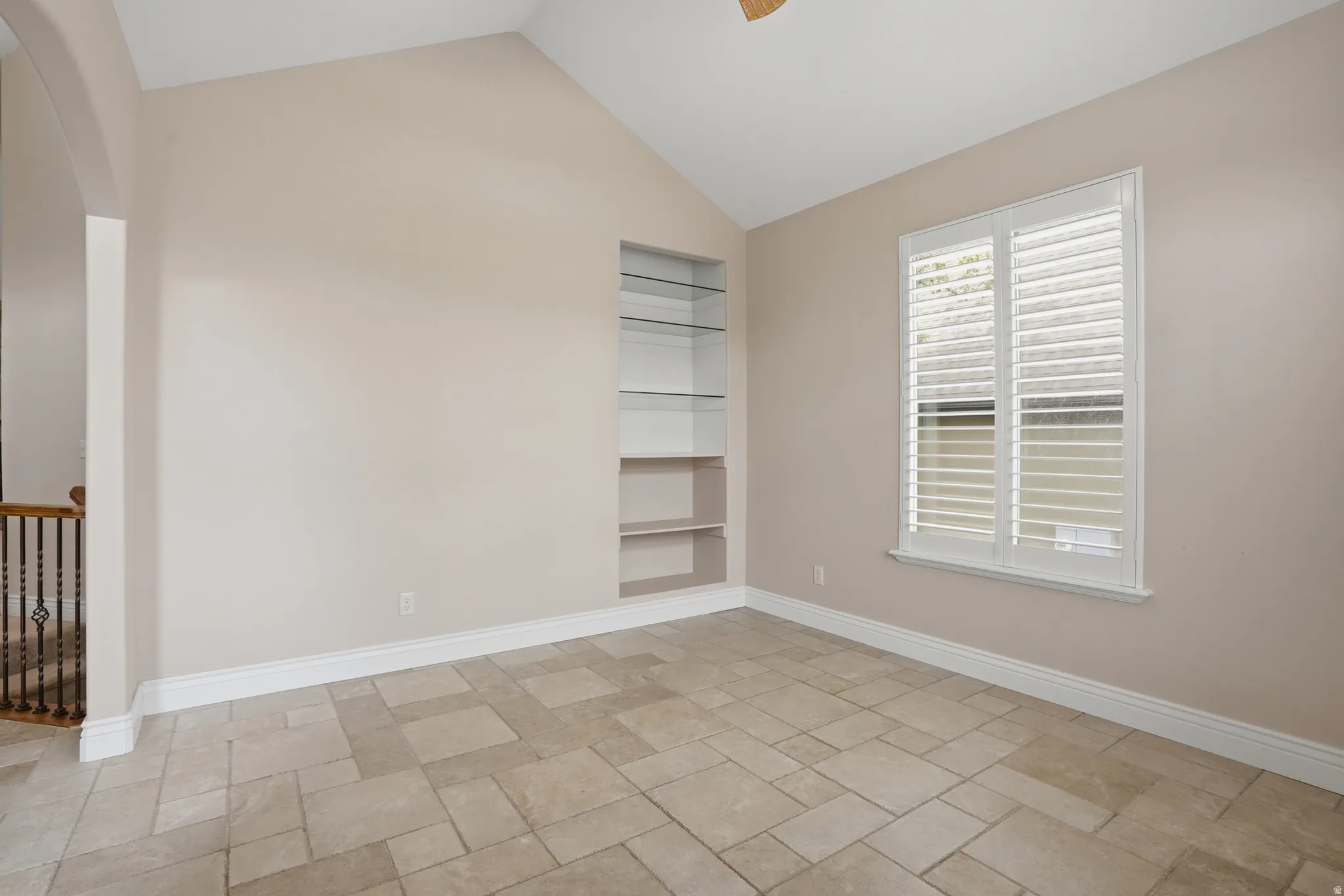 Vaulted ceiling, built-in shelving, window with plantation shutters, arched doorway, and tile flooring