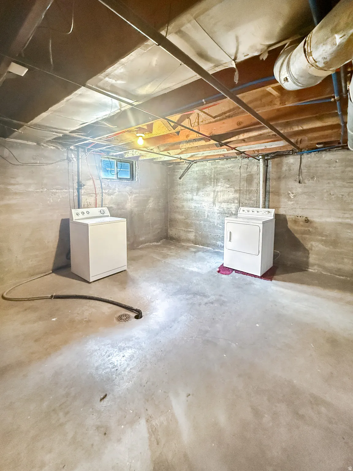 Concrete flooring in a spacious utility area featuring exposed joist ceiling, a utility sink drain, and a small window
