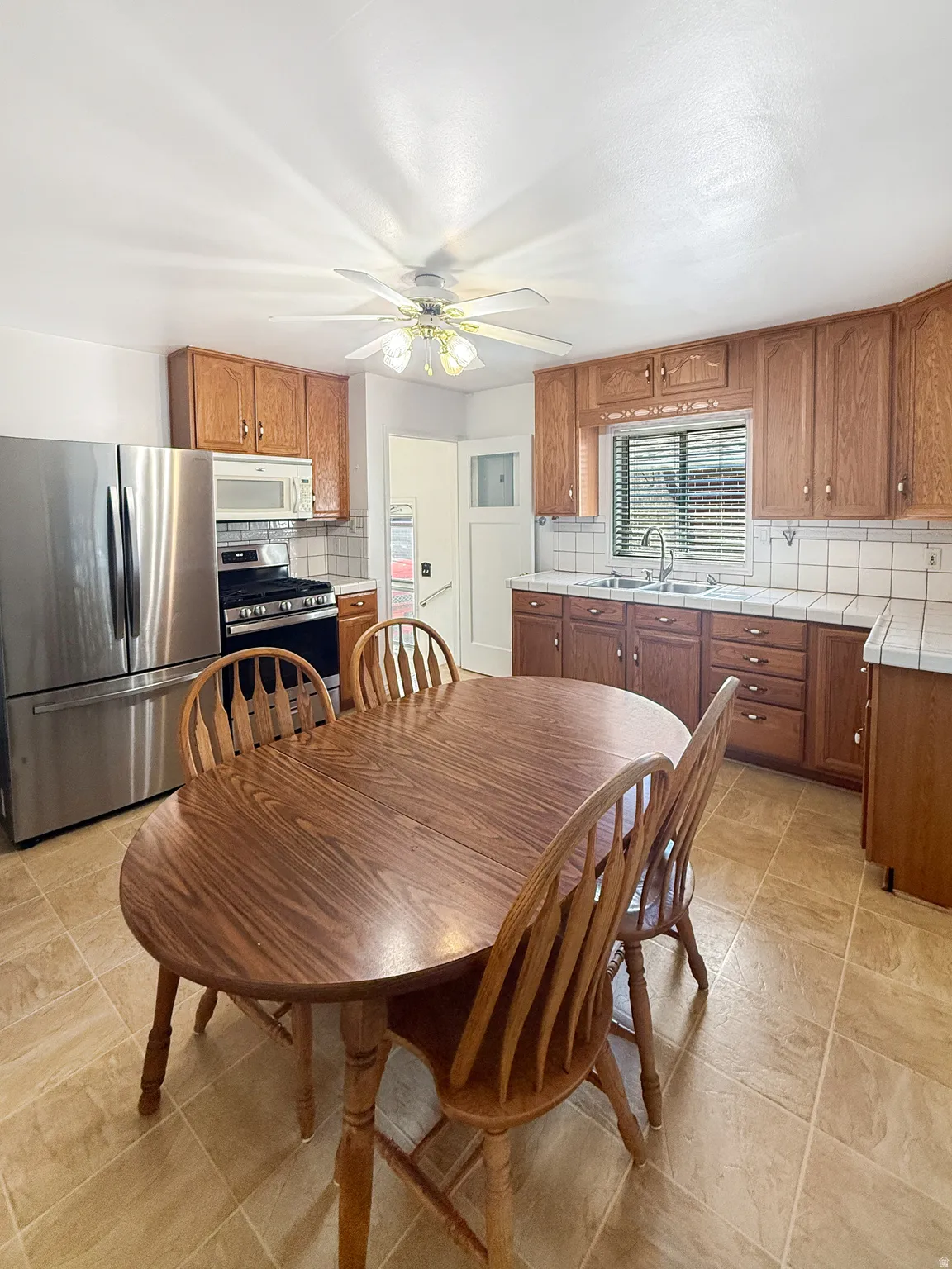 Kitchen featuring wood cabinetry, stainless steel appliances, a double basin sink with a gooseneck faucet, light-toned tiled flooring, and a ceiling fan with integrated lighting