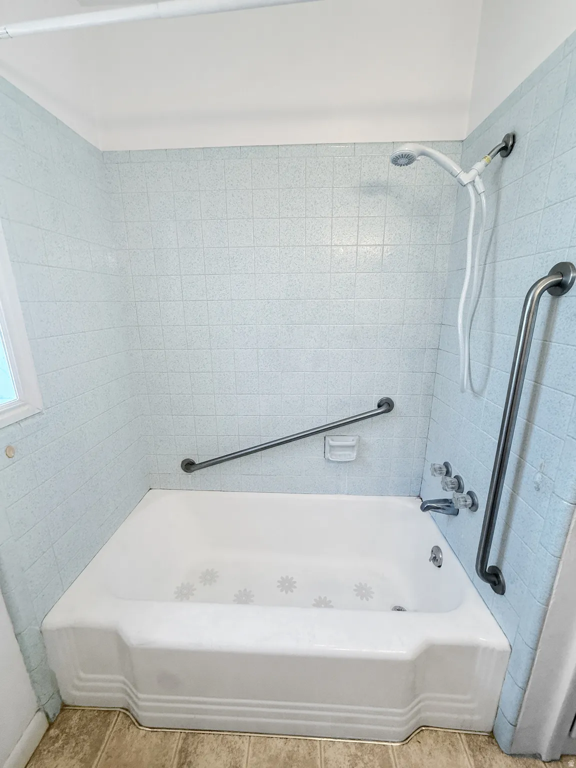 Bathtub with light blue tiled surround featuring a handheld showerhead, chrome fixtures, and two grab bars