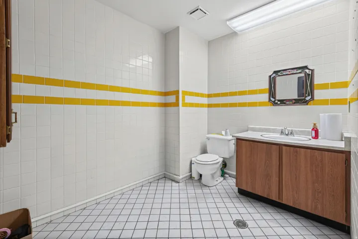 White and yellow accent wall tile, white floor tile, wood-finish vanity with a single sink, white countertop, and fluorescent overhead lighting