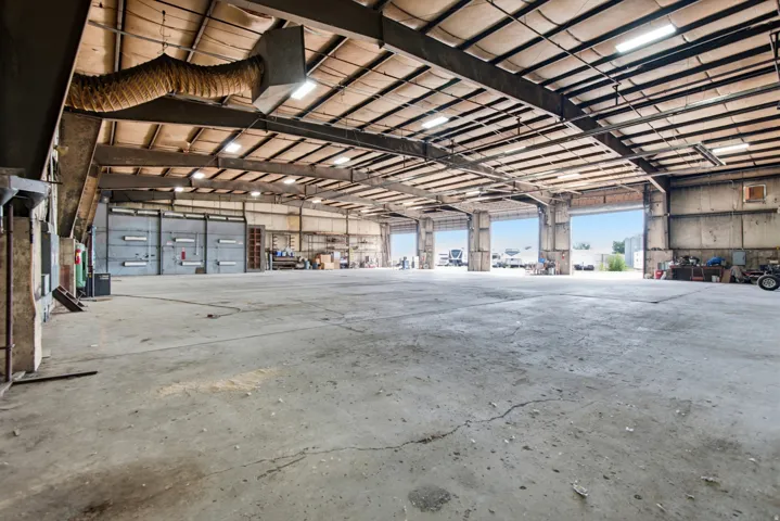 Expansive concrete flooring with multiple roll-up doors, exposed metal beam ceiling, and fluorescent lighting fixtures