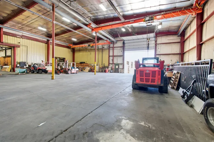 Expansive concrete flooring with high ceilings, featuring exposed structural beams, overhead gantry cranes, and a large roll-up door