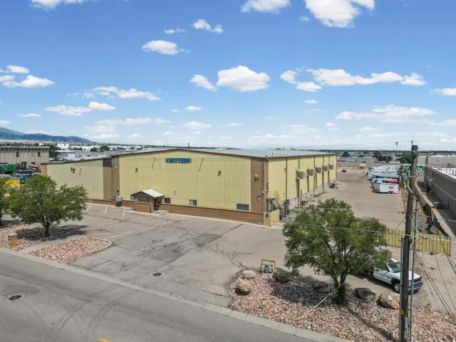 Industrial warehouse featuring a corrugated metal exterior with yellow finish, expansive paved lot with ample parking, mature landscaping with established trees, secure perimeter fencing, and clear sightlines across the property with mountain backdrop vis