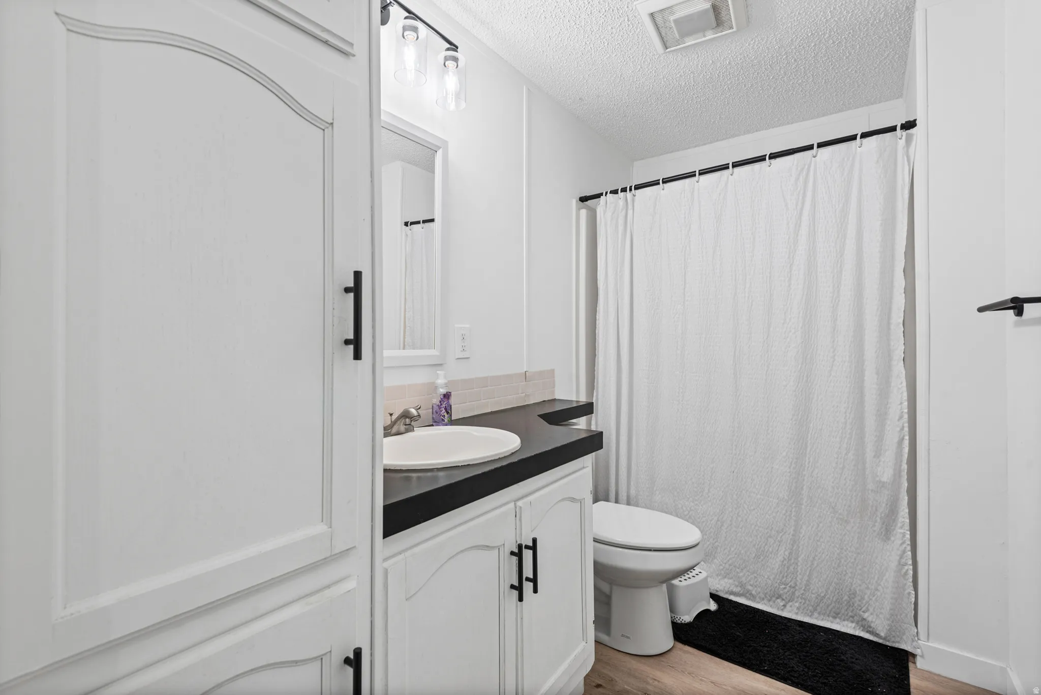 White cabinetry with black hardware, dark countertop, single basin sink, wall-mounted mirror, and wood-finish flooring
