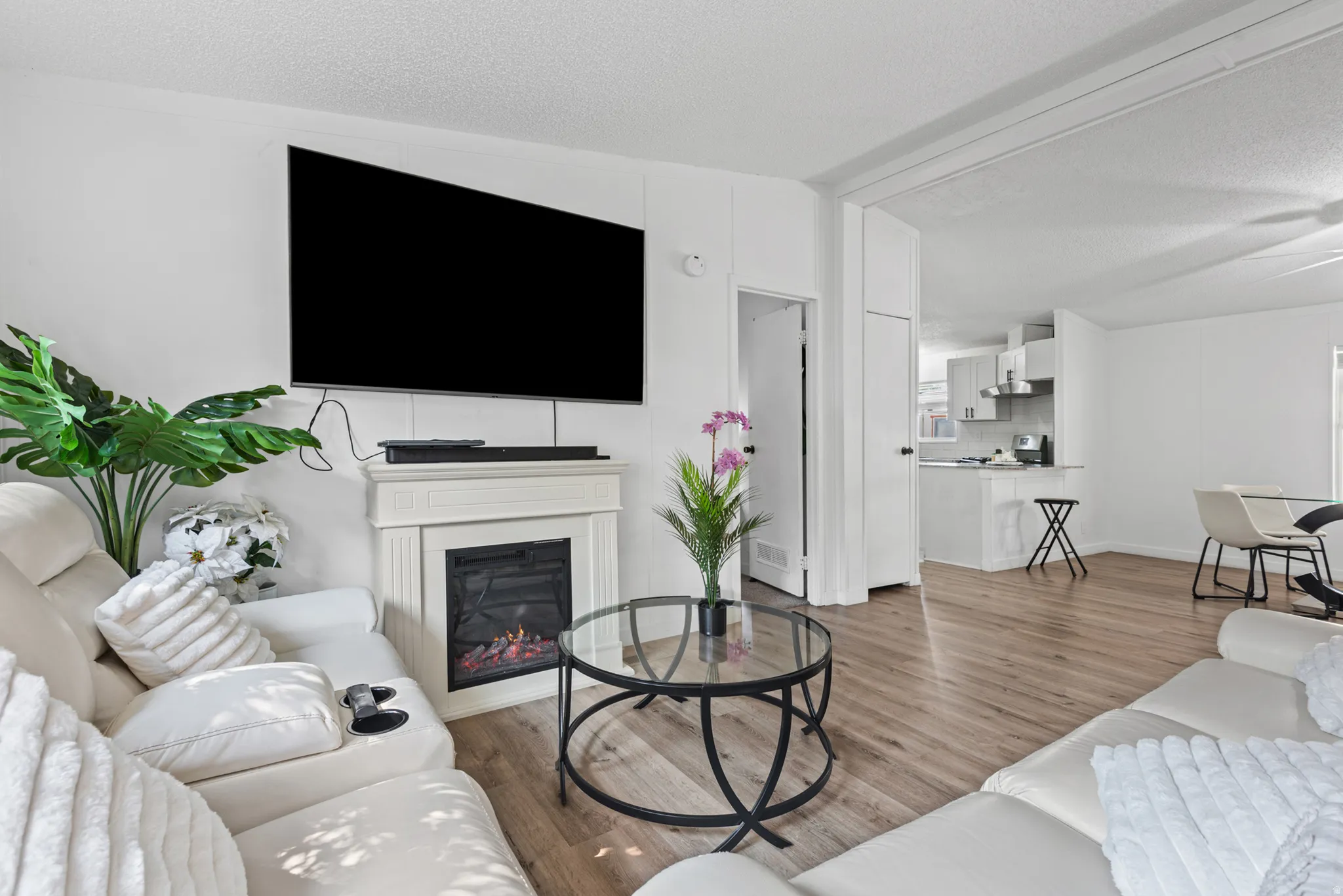 Wood-finish flooring throughout, featuring a white fireplace surround with an electric insert, and an open doorway leading to a kitchen with white cabinetry