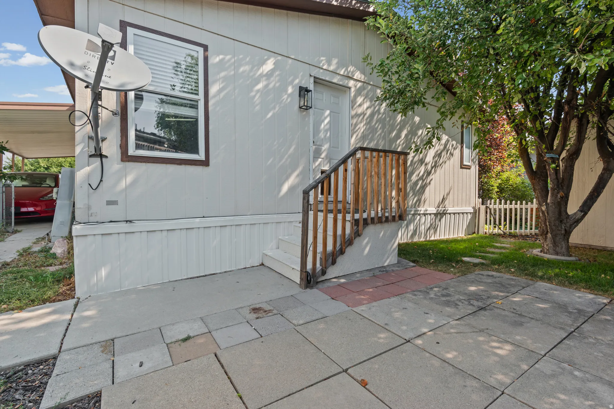 White siding exterior with a wood-stained entry railing, concrete patio, and mature tree
