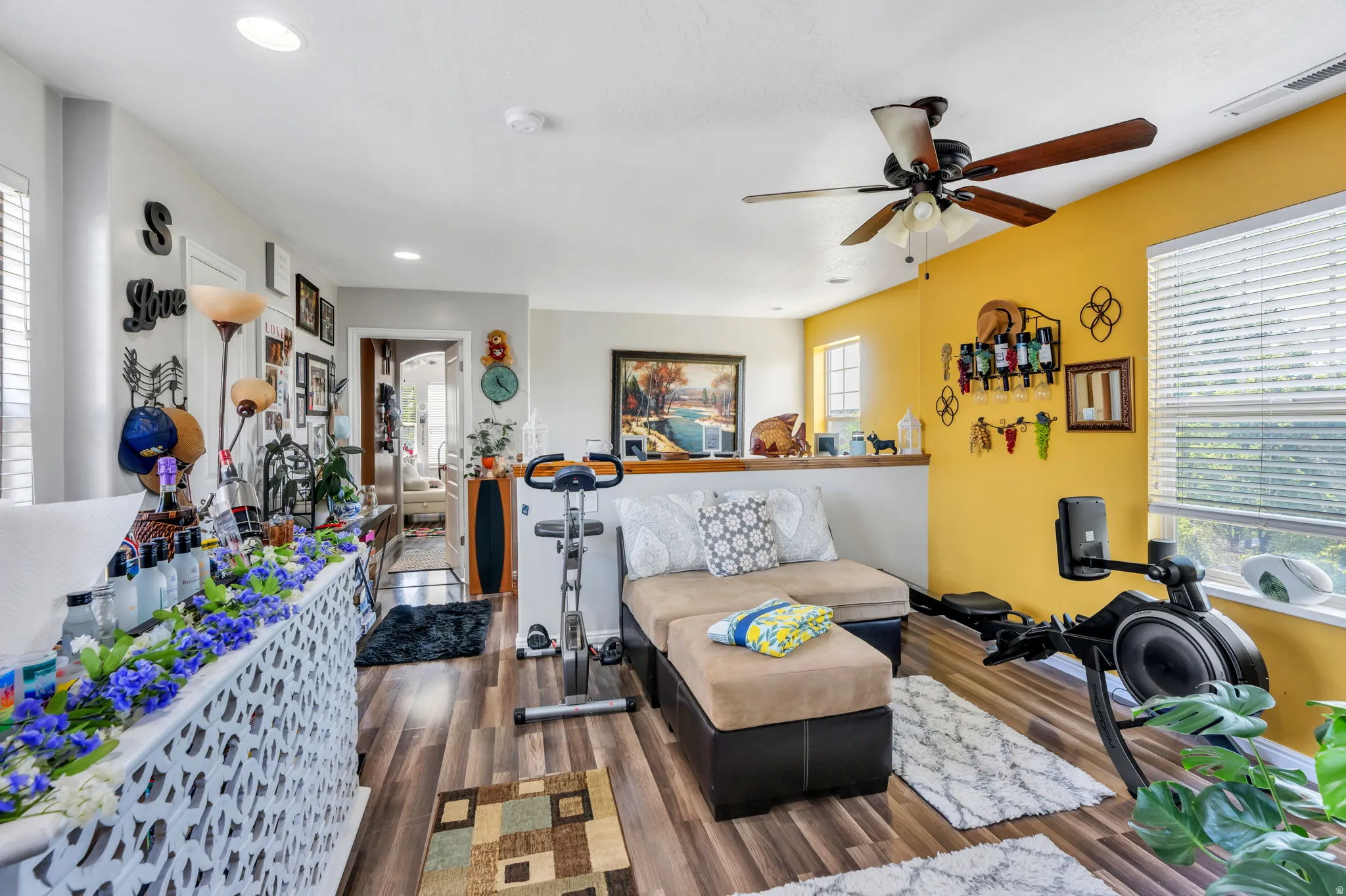Bright, open living space featuring warm yellow accent walls, wood-finish flooring throughout, ceiling fan with integrated lighting, and built-in wall-mounted storage and shelving