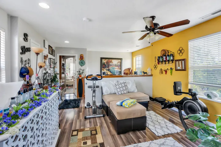 Bright, open living space featuring warm yellow accent walls, wood-finish flooring throughout, ceiling fan with integrated lighting, and built-in wall-mounted storage and shelving