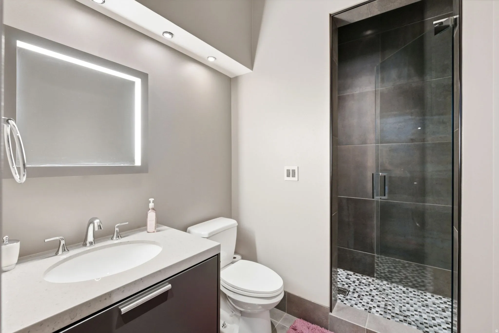 Vanity with integrated sink and chrome fixtures, illuminated mirror with backlighting, recessed ceiling lights, glass-enclosed shower with large format tile surround, and pebble-finish shower floor