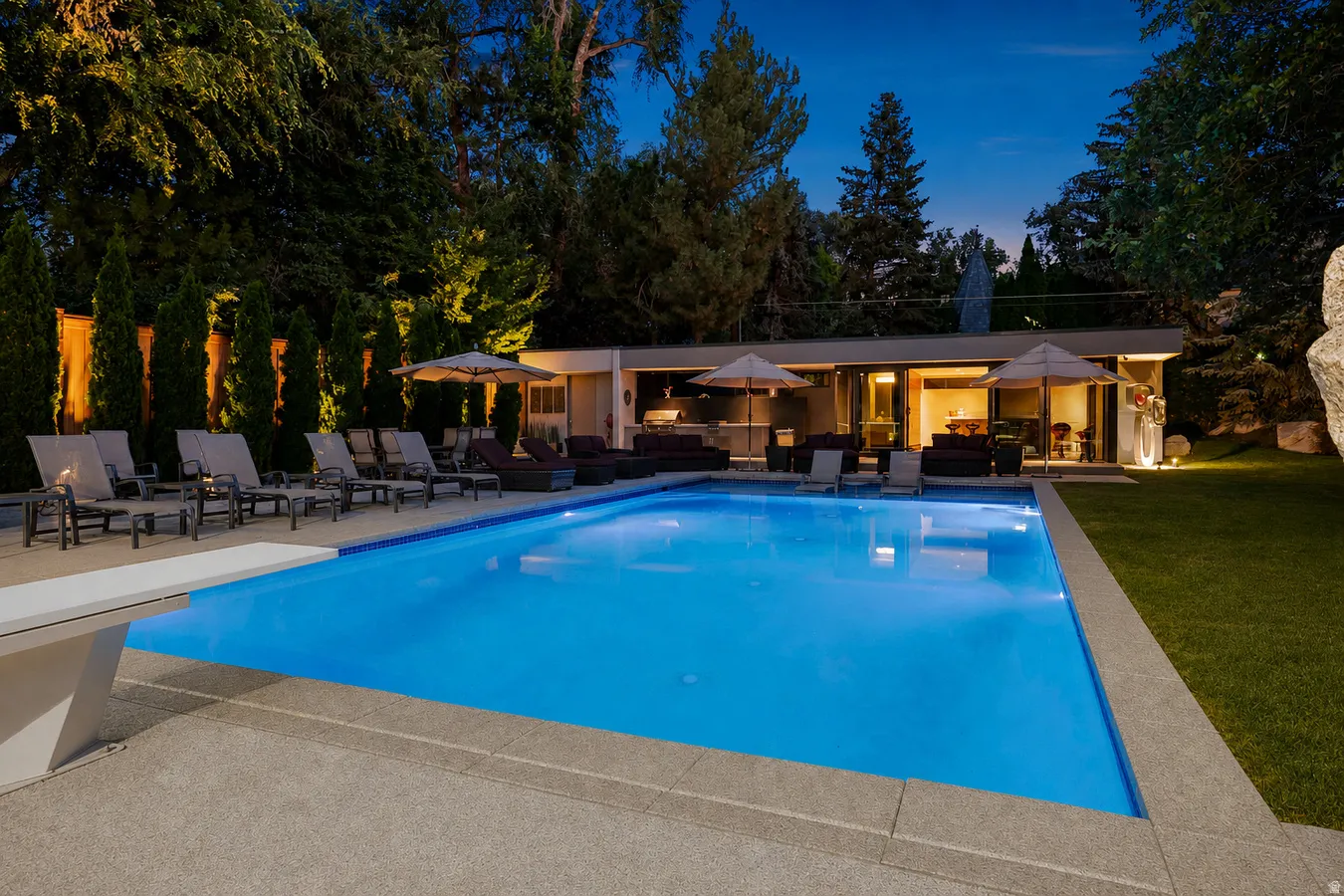 In-ground swimming pool with diving board, expansive paved patio, lush lawn, and a modern cabana