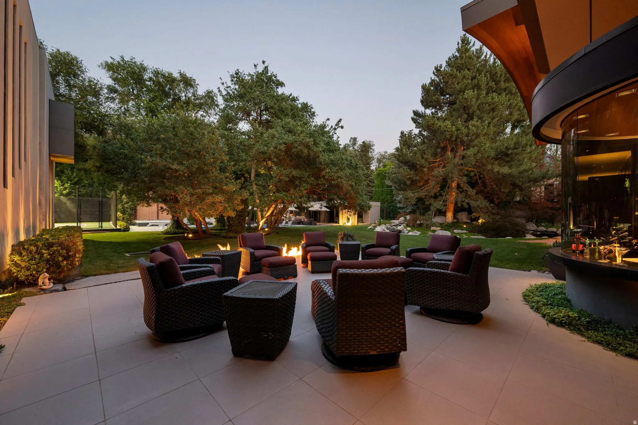 Expansive paved patio featuring a built-in fire pit, surrounding lawn, and mature trees