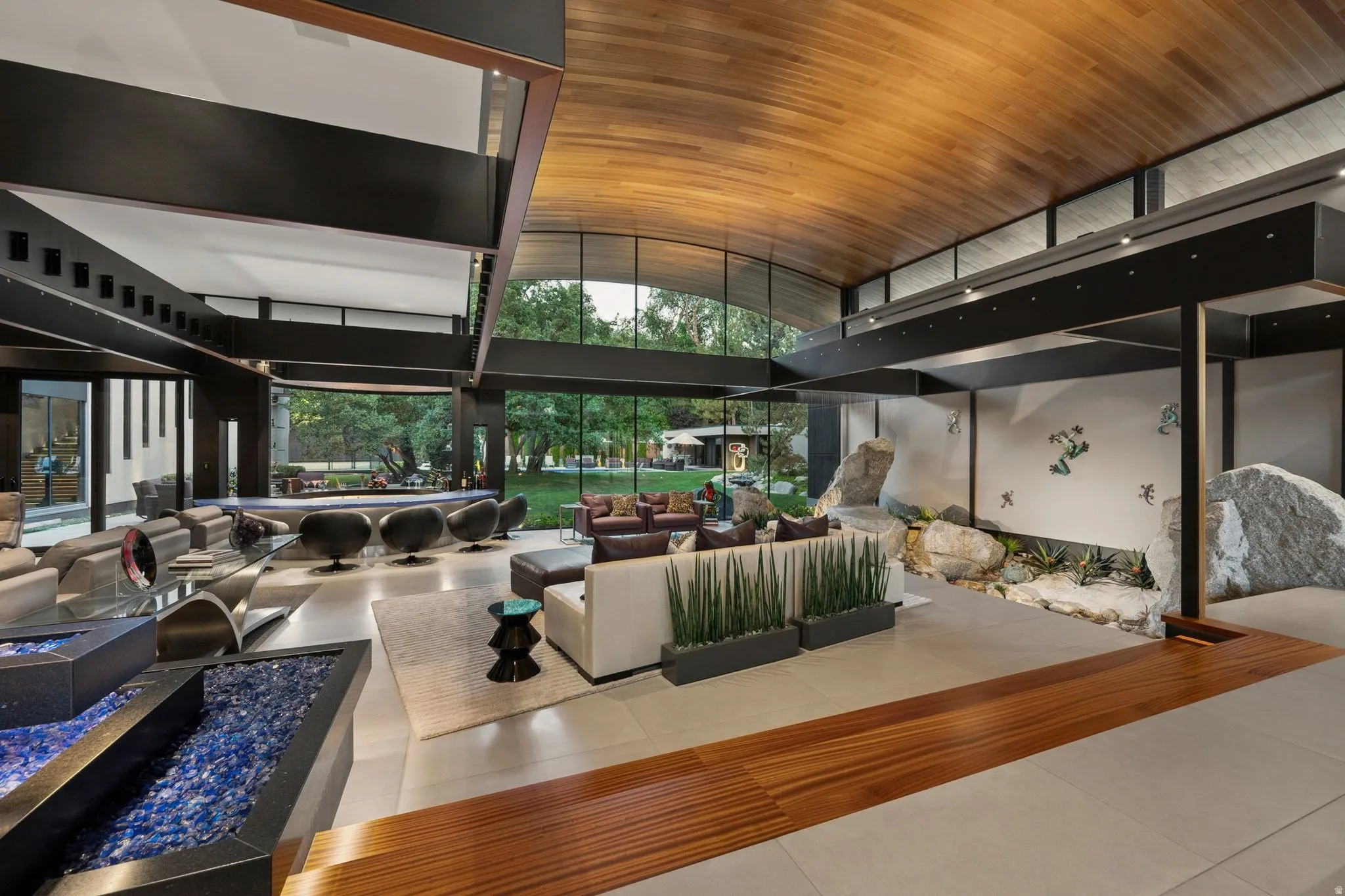 Vaulted wood-paneled ceiling, expansive window walls, built-in indoor rock garden, multi-tiered water feature with blue glass, and tile flooring with wood-finish accents