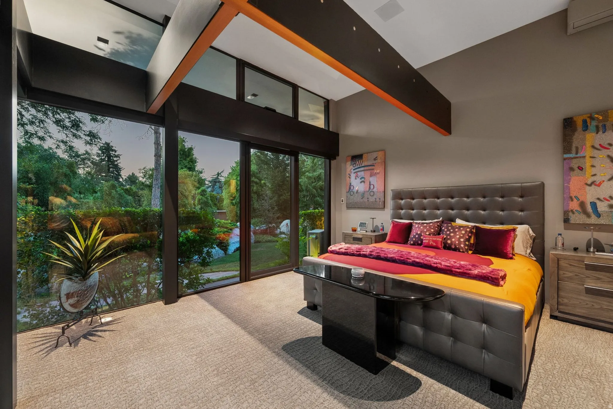 Floor-to-ceiling windows, vaulted ceilings with wood beams, light gray carpeting, and a tufted bed frame with an upholstered headboard