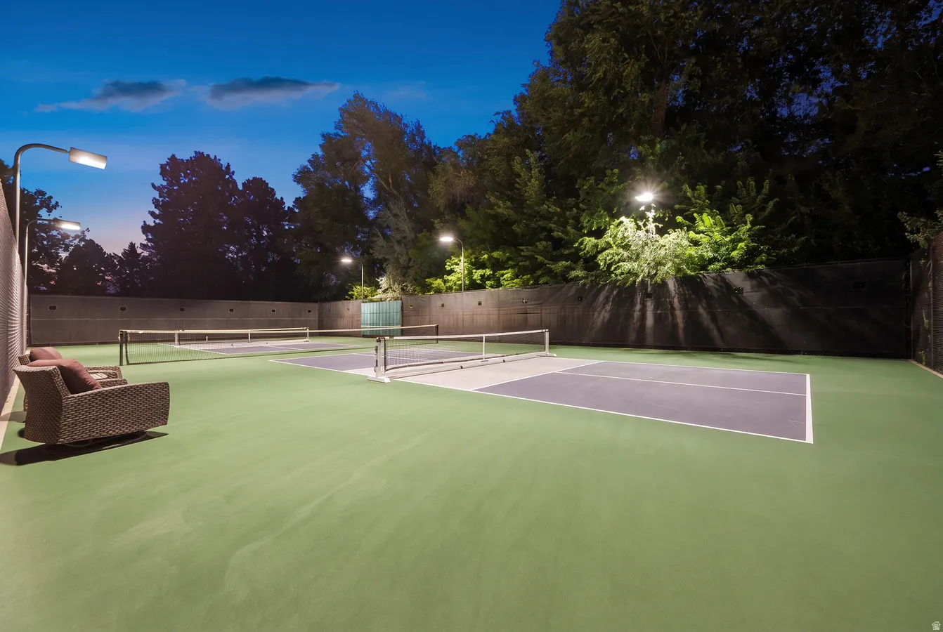Two pickleball courts featuring a green and gray surface, perimeter fencing, and overhead lighting