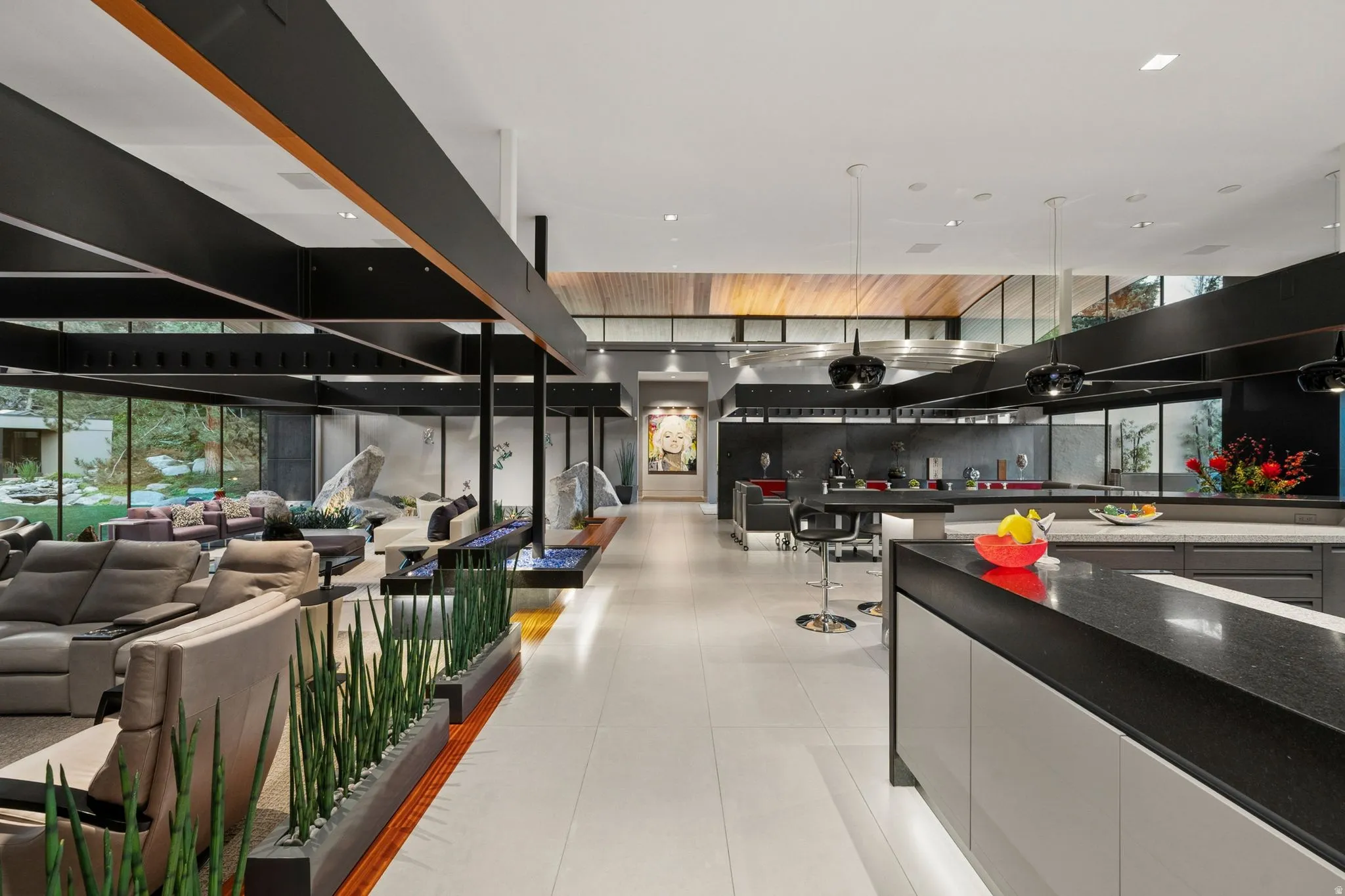 Open-concept interior featuring high ceilings, exposed structural beams, a linear fireplace with blue fire glass, a kitchen island with dark countertops, and extensive perimeter glass panels
