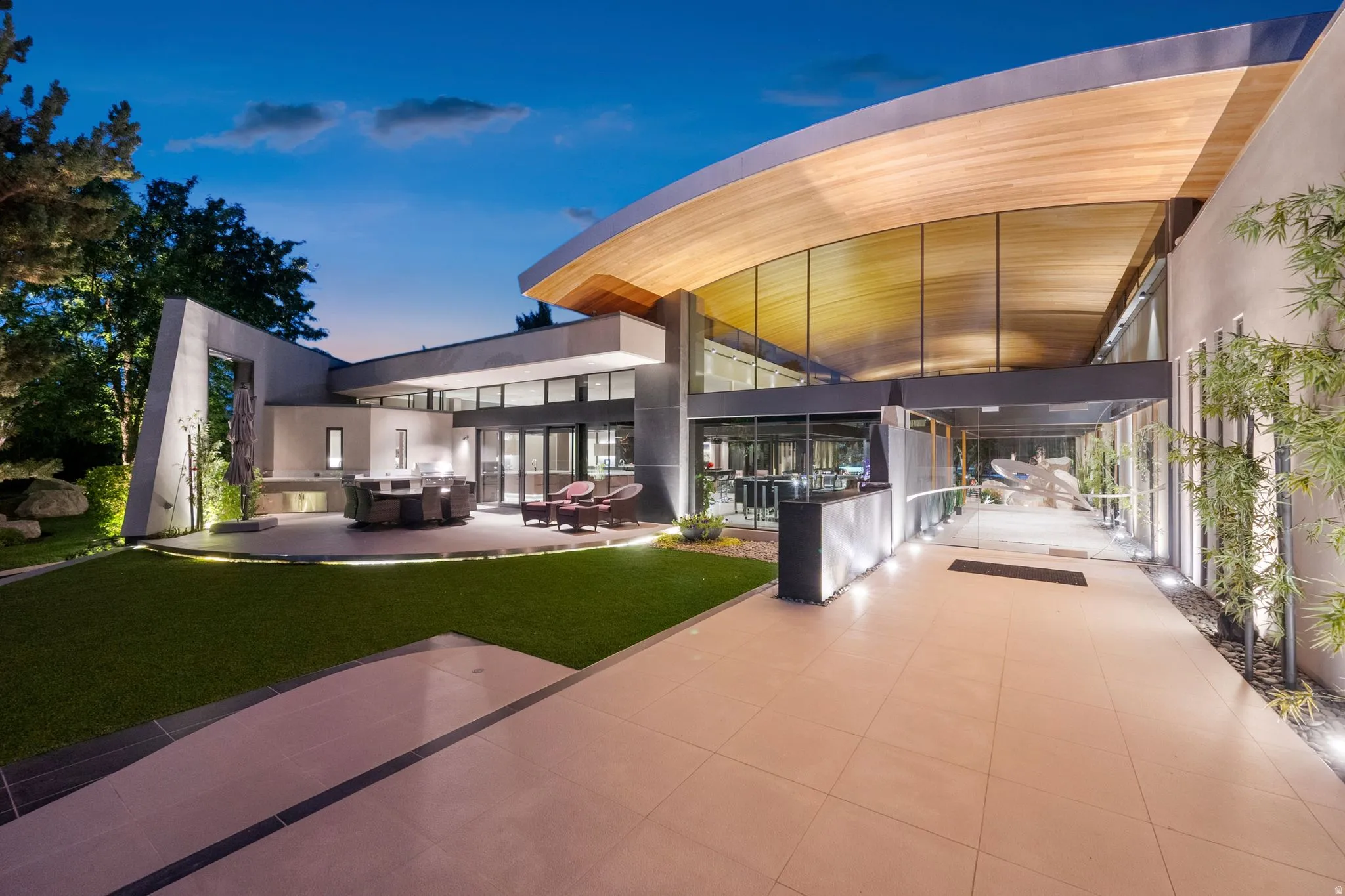Curved architectural roofline with wood-finish soffit, expansive glass facade, tiled outdoor patio, manicured lawn, and dedicated pathway lighting