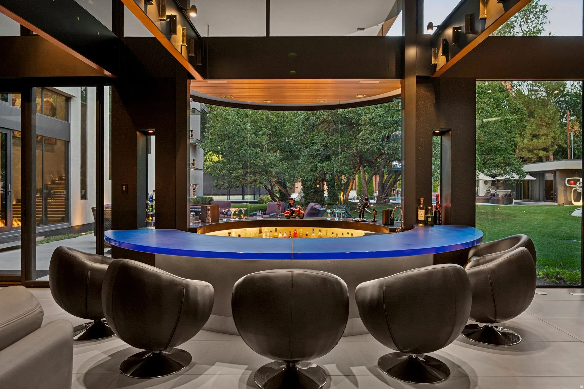 Indoor-outdoor living space featuring a curved wet bar with a blue countertop, expansive full-height windows, structural wood beam accents, and a built-in outdoor seating area