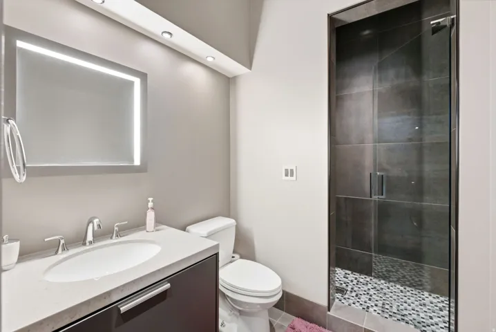 Vanity with integrated sink and chrome fixtures, illuminated mirror with backlighting, recessed ceiling lights, glass-enclosed shower with large format tile surround, and pebble-finish shower floor
