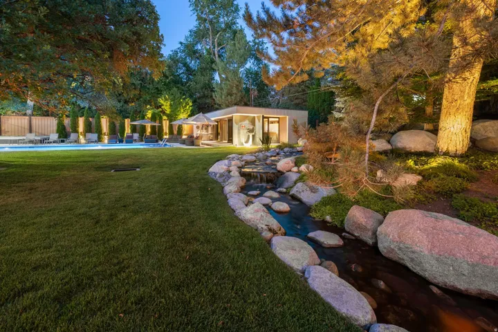 Expansive backyard featuring a large swimming pool, lush green lawn, natural rock stream with a waterfall, mature trees, and a modern accessory structure