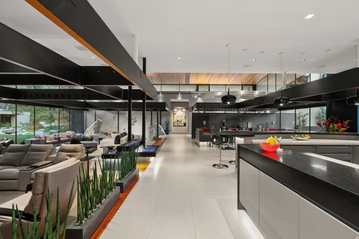 Open-concept interior featuring high ceilings, exposed structural beams, a linear fireplace with blue fire glass, a kitchen island with dark countertops, and extensive perimeter glass panels
