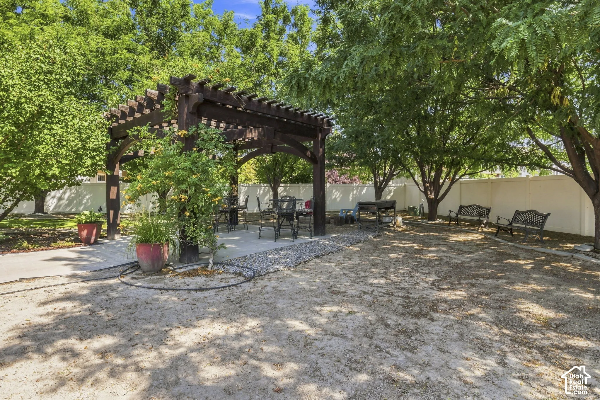 Spacious outdoor area featuring a dark wood pergola over a paved patio, gravel ground cover, and mature trees