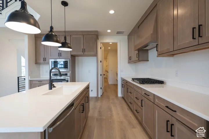Modern kitchen featuring a large island with an undermount sink, light-toned wood-finish flooring, recessed lighting, black industrial pendant fixtures, and stainless steel appliances