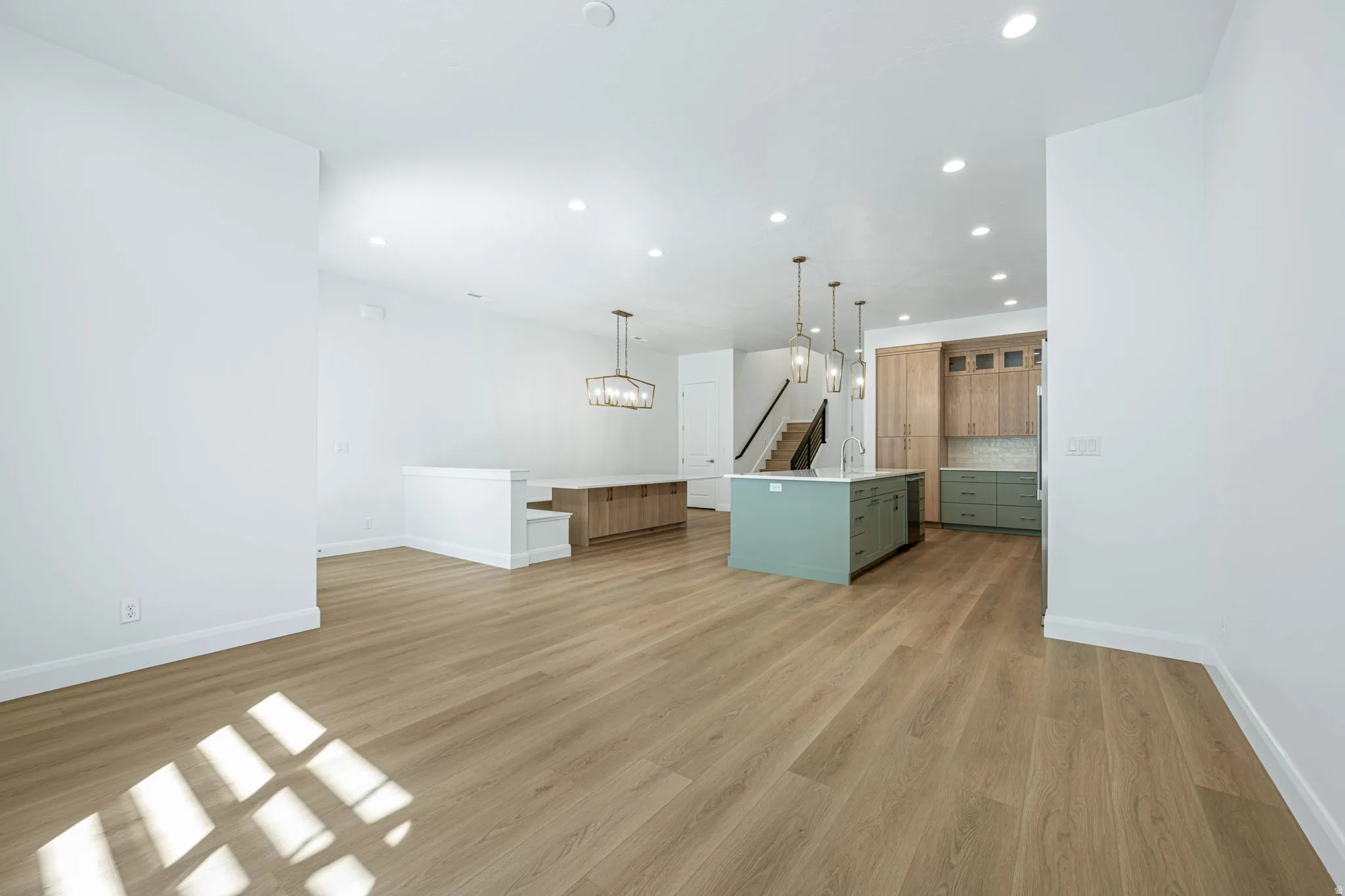 Wood-finish flooring throughout, recessed ceiling lighting, kitchen island with pendant lighting, light wood-finish cabinetry, and a subway tile backsplash