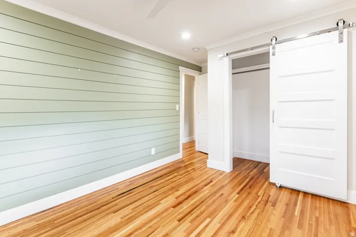 Spacious bedroom featuring soft green shiplap accent wall, warm wood-finish flooring throughout, and a modern sliding barn door entry