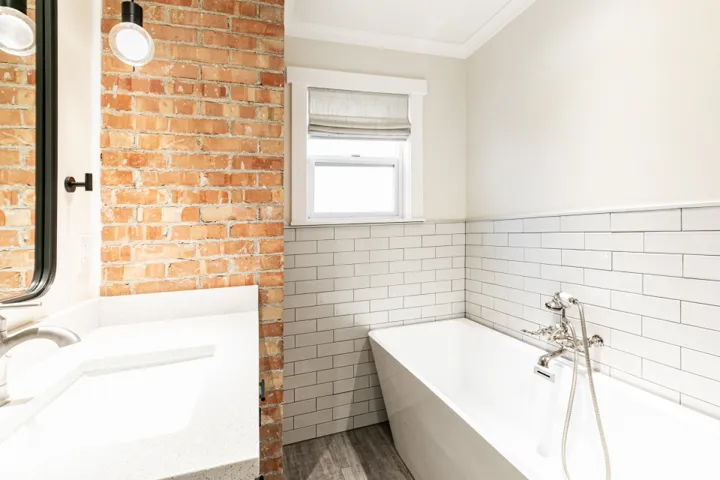 Modern bathroom featuring exposed brick accent wall, white subway tile surround, freestanding soaking tub with deck-mounted faucet, floating vanity with integrated sink, and natural light from casement window