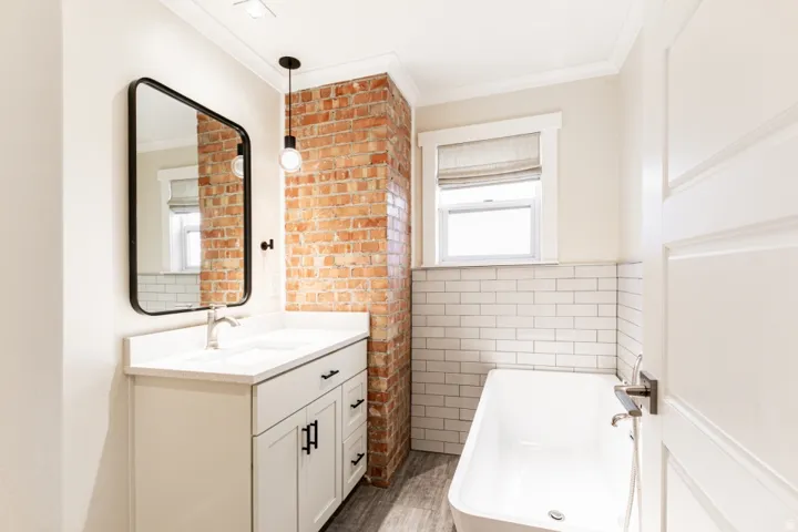 White vanity with dual integrated sinks, subway tile wainscoting, exposed brick accent wall, and a standalone soaking tub