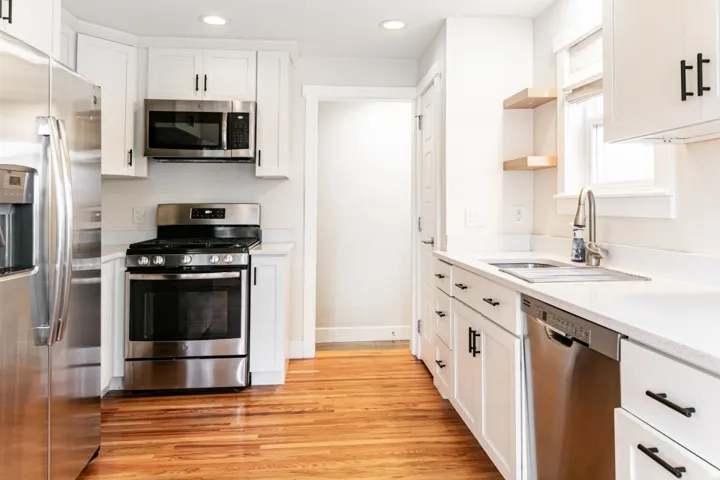 White cabinetry with black hardware, stainless steel appliances, light-toned countertops, wood-finish flooring, and open shelving
