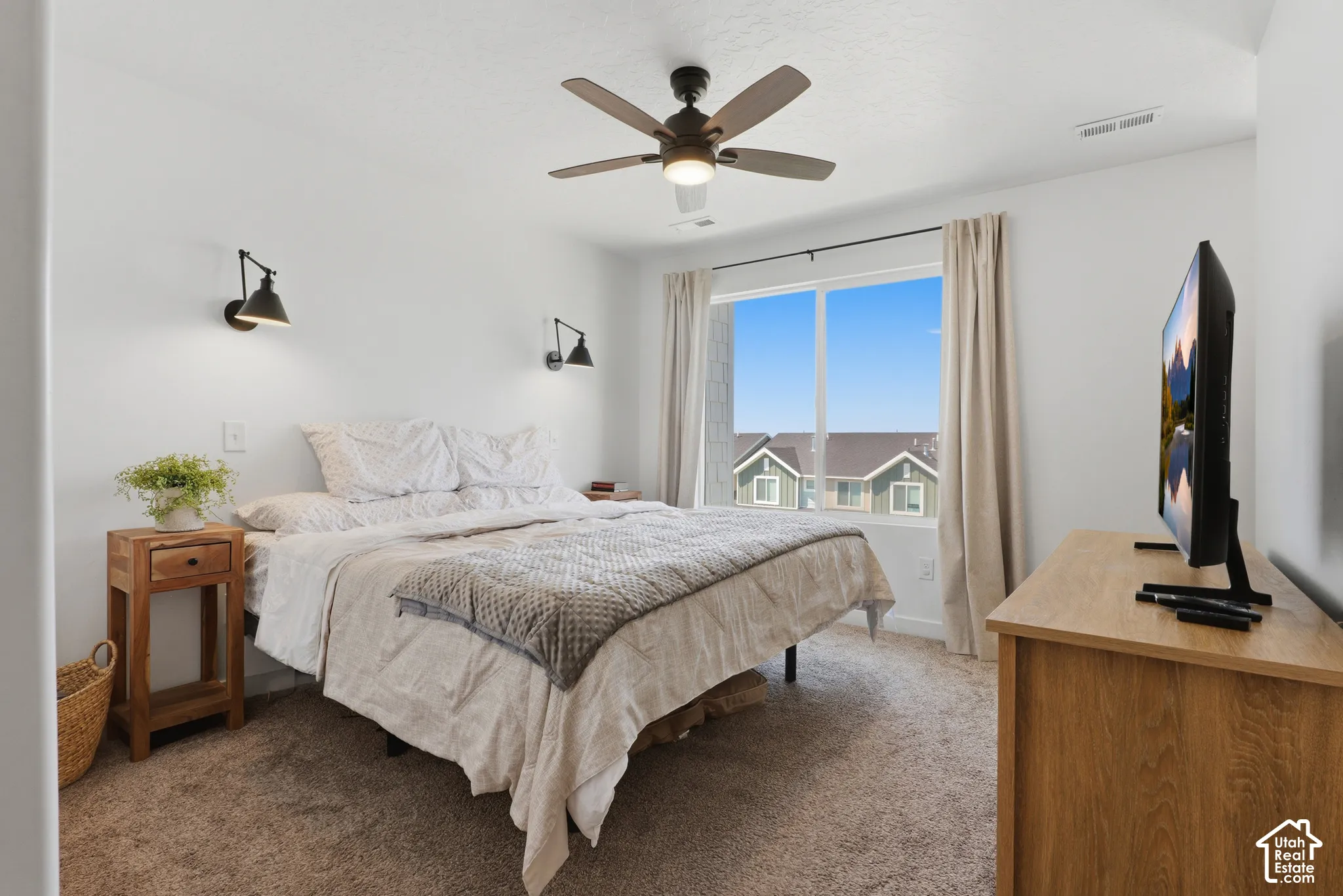 Wall-mounted sconce lighting, ceiling fan with integrated light, large window with roller shades, and textured carpeting