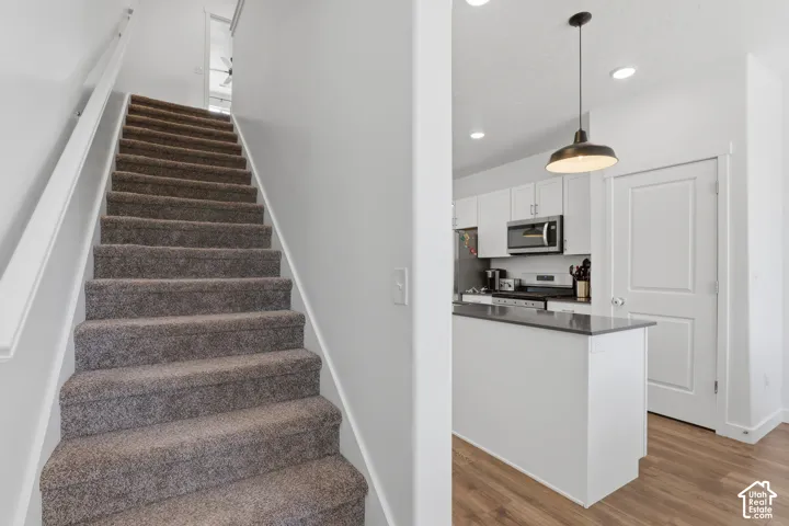 Carpeted staircase with white handrail, recessed lighting, wood-finish flooring, and a kitchen island with a dark countertop