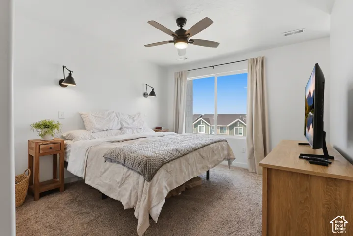 Wall-mounted sconce lighting, ceiling fan with integrated light, large window with roller shades, and textured carpeting
